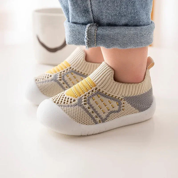 A pair of beige and yellow breathable mesh baby shoes resting on a white surface next to denim pants.