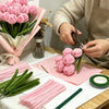 A person is preparing a DIY pipe cleaner flower bouquet using pink and green pipe cleaners on a wooden table.