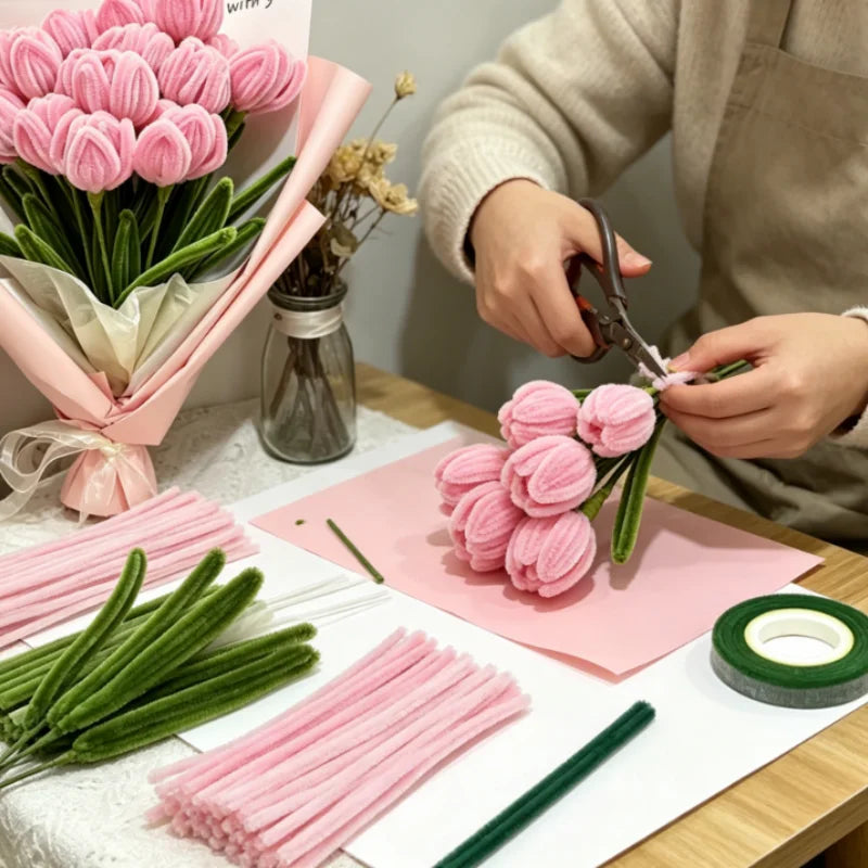 A person is preparing a DIY pipe cleaner flower bouquet using pink and green pipe cleaners on a wooden table.
