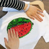 A hand is holding a green sequin watermelon design on a white t-shirt.