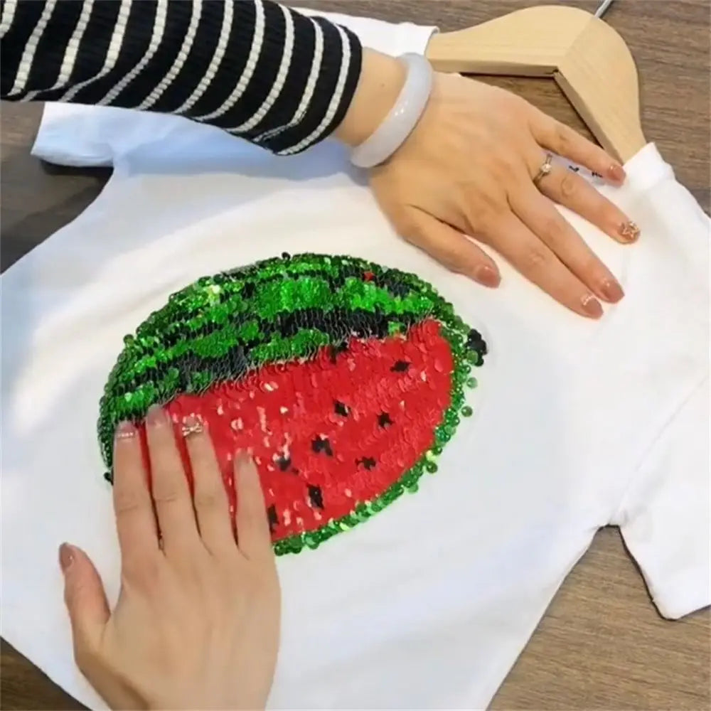 A hand is holding a green sequin watermelon design on a white t-shirt.