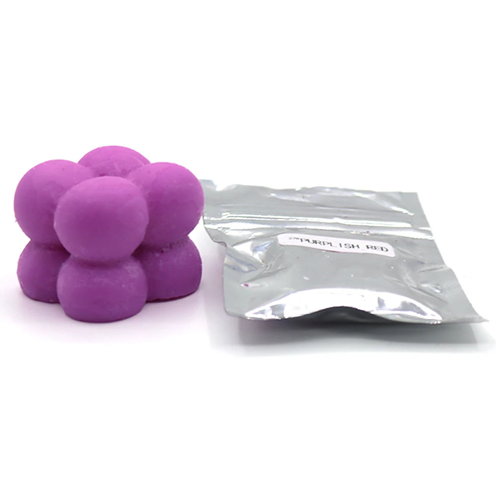 A pile of purple candle dye chips next to a small silver plastic bag labeled purple ish red.