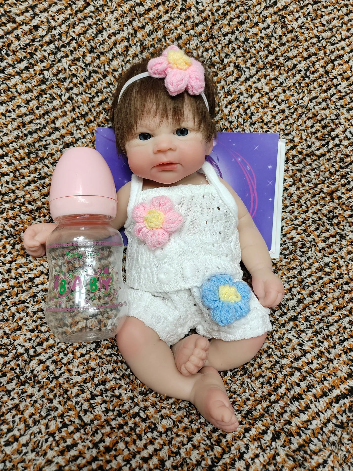 A realistic soft silicone reborn doll with pink hair, floral accessories, and a purple book lying next to it.
