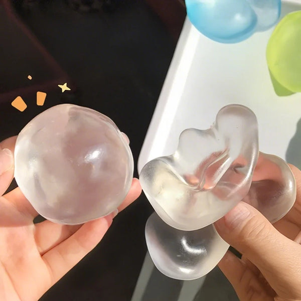 Une main tient un jouet anti-stress en huile de coco rose translucide en forme de cœur, à côté de ballons bleus et verts.