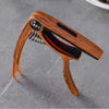 A wooden guitar capo with a metal pick holder and red grip is shown on a gray surface.