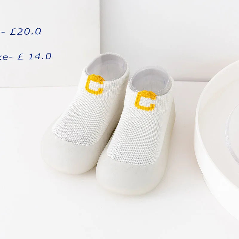 A pair of white knitted soft sole sock shoes for babies and toddlers with yellow 'C' emblems.