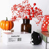 A handmade crocheted bat doll in black is displayed next to a glass jar and red floral branches on a white background.