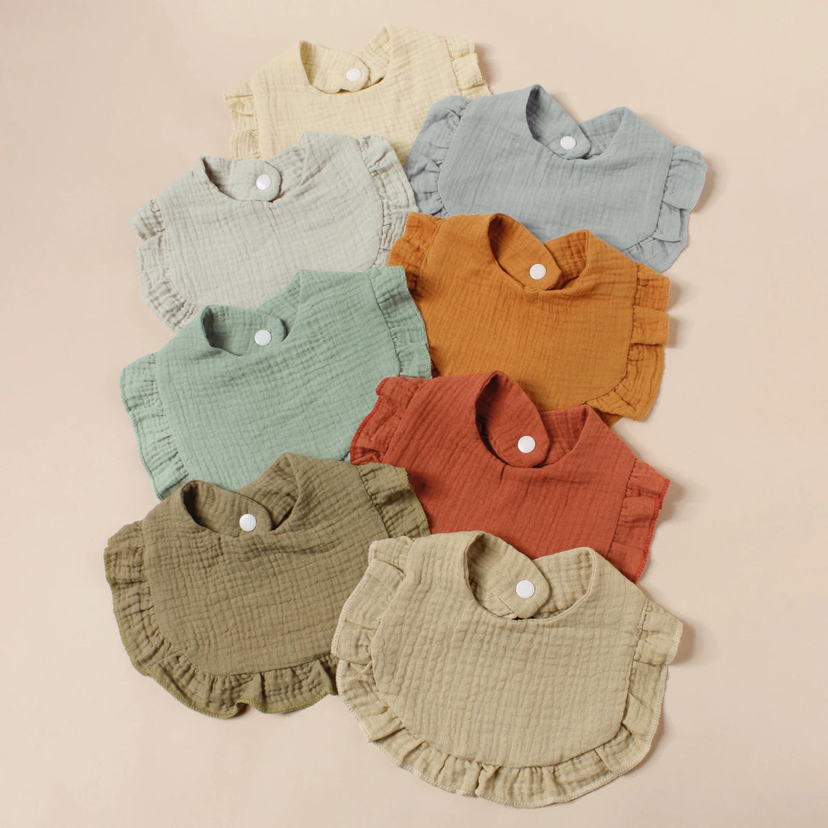 A set of eight cotton ruffle baby bibs in various muted earth tones including beige, sage green, mustard yellow, and terracot