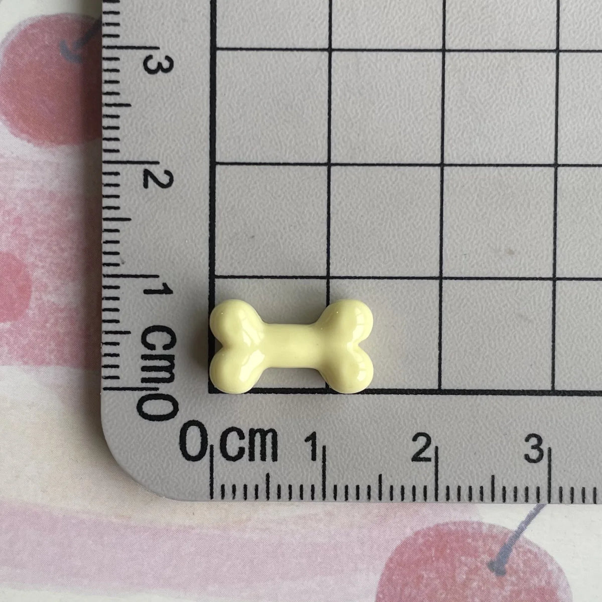 A small, pale yellow resin bone charm is placed on a ruler marked in centimeters.