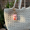 A handmade crochet mini pink pig keychain hanging from a gold-colored loop on a light beige woven bag.