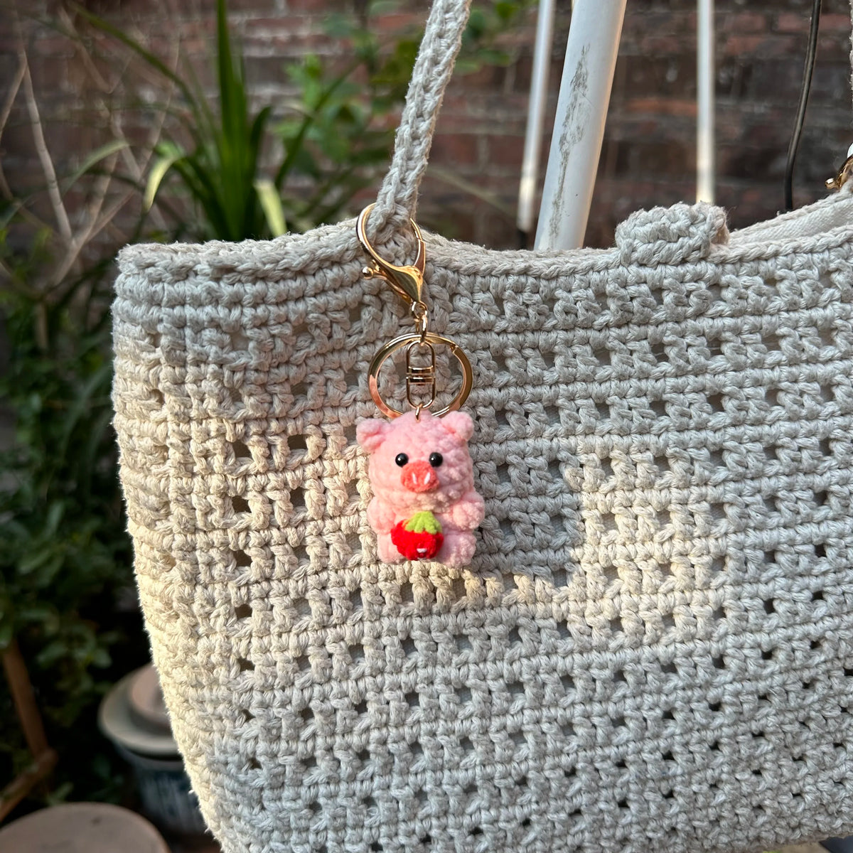 A handmade crochet mini pink pig keychain hanging from a gold-colored loop on a light beige woven bag.