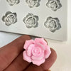 A hand holds a pink rose flower silicone mold for candle and soap making next to a white silicone tray.