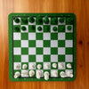 A green chessboard with white squares and 32 light-colored chess pieces arranged on the board.