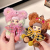 A hand holds a pink and green fluffy plush charm featuring a face with pink hair and a plaid outfit.