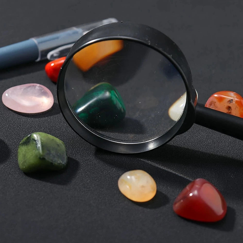 A magnifying glass shows various colored natural crystals and minerals on a dark surface.
