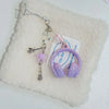 A beaded wing cross phone lanyard keychain with a purple earbud resting on a white fluffy surface.