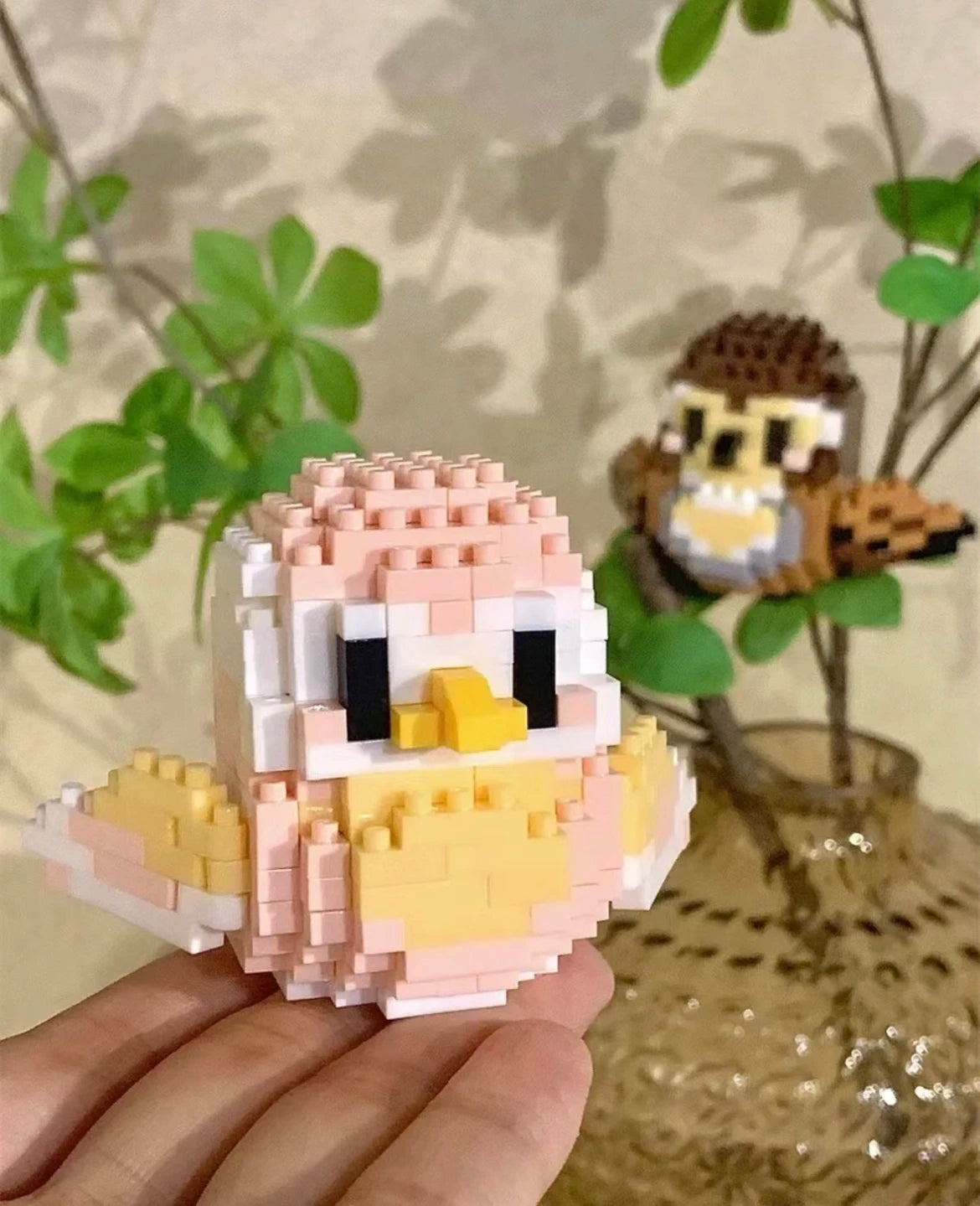 A hand holds a colorful parrot micro building blocks set featuring pink, yellow, and white pieces against a blurred green bac