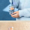 A small, gray teddy bear is attached to a phone strap and held by a person against a blue background.