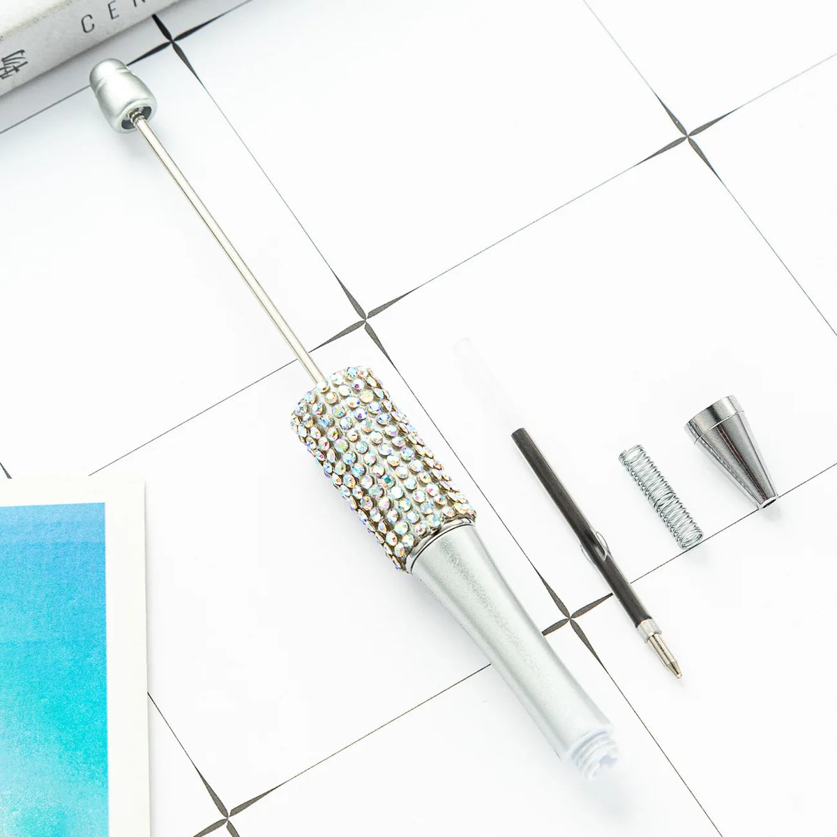 A silver ballpoint pen craft kit featuring a beaded tip and various tools on a white surface.