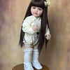 A lifelike 55 cm soft silicone baby doll with long dark hair and a white outfit standing on a wooden surface.