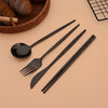 A set of six black stainless steel Korean dinnerware including a spoon, fork, and chopsticks resting on a light brown surface