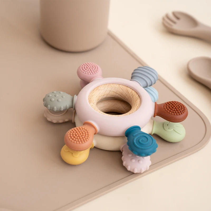 A set of colorful rudder-shaped silicone baby teethers with wooden rings arranged on a light beige tray.