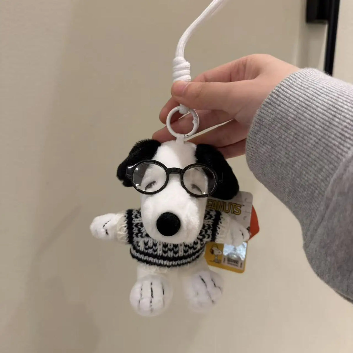 A hand is holding a white dog plush keychain charm with black ears and round glasses against a light wall.