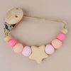 A wooden pacifier clip with silicone beads in pink and peach colors on a light beige background.