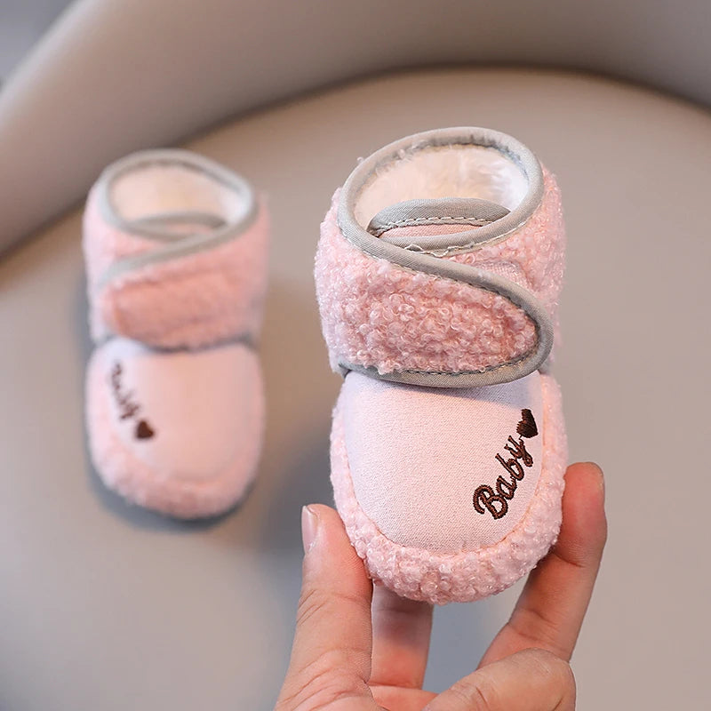 A hand holds a pair of pink plush baby snow boots with gray trim and anti-slip soles.