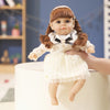 A lifelike 14-inch reborn baby doll with a brown braided hairstyle and white lace outfit is being held by a hand.