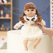 A lifelike 14-inch reborn baby doll with a brown braided hairstyle and white lace outfit is being held by a hand.