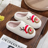 A pair of white plush indoor slippers with reindeer and Santa designs resting on a wooden tray.