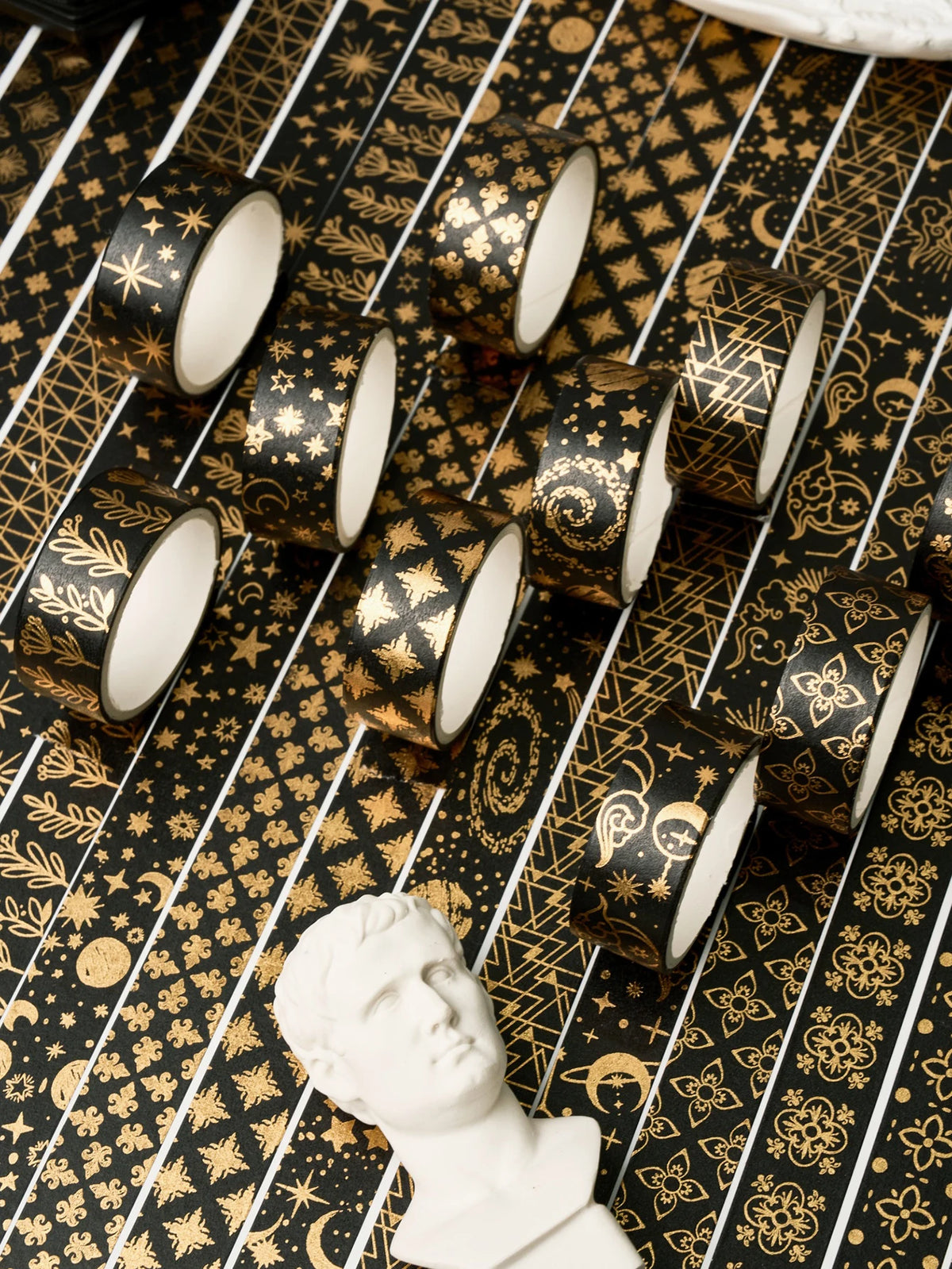 A collection of black and gold foil washi tape rolls scattered on a patterned surface next to a white bust.