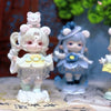 A white collectible figurine of a girl with pink ears and blonde hair wearing a yellow dress and a blue hat.