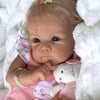 A close-up of an 18-inch lifelike reborn baby doll with hand-rooted hair wearing a pink dress and holding a white plush toy.