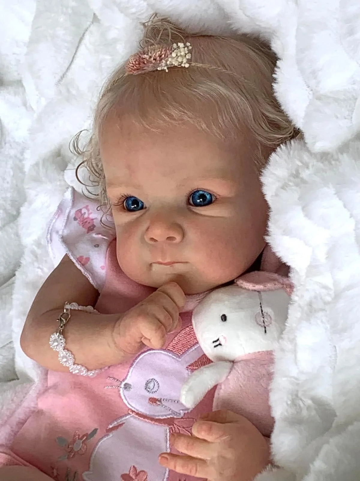 A close-up of an 18-inch lifelike reborn baby doll with hand-rooted hair wearing a pink dress and holding a white plush toy.