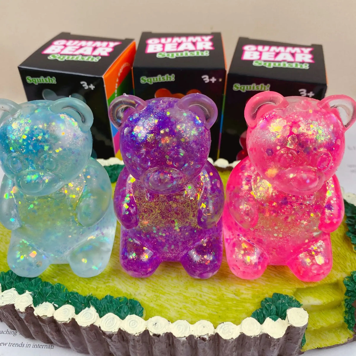 A display of three gummy bear squishy toys in purple, pink, and green packaging on a yellow cake.