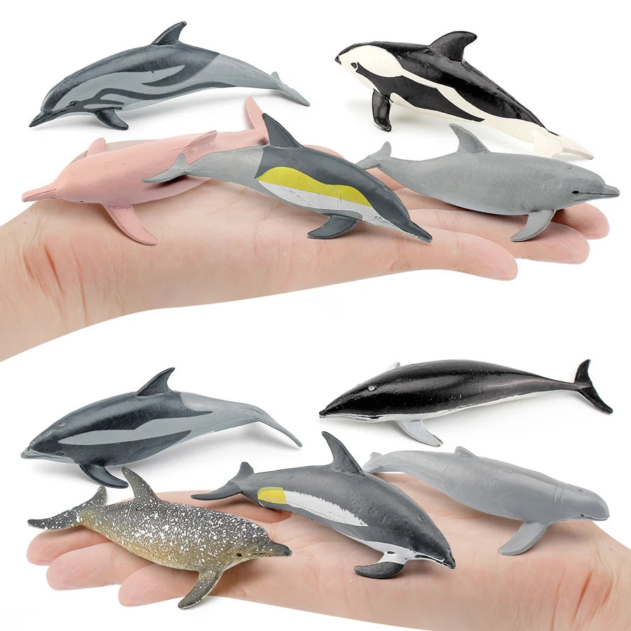 A hand holds a set of realistic mini ocean animal figurines including a whale, shark, dolphin, turtle, and penguin.