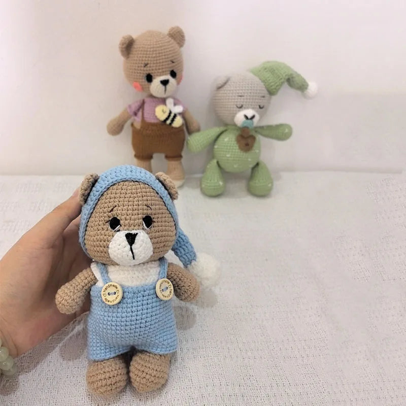 A hand holds a handmade crochet stuffed bear in light blue overalls and brown fur.