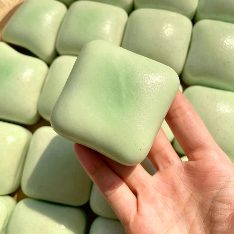 A hand holds a light green sticky cube slow-rising squishy stress relief toy surrounded by many similar cubes.