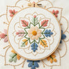 A white circular embroidery hoop featuring a detailed floral mandala design in pink, green, blue, and yellow thread.