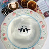 A white squishy toy shaped like a cat face is placed in a decorative plate surrounded by small pink flowers and strawberries.
