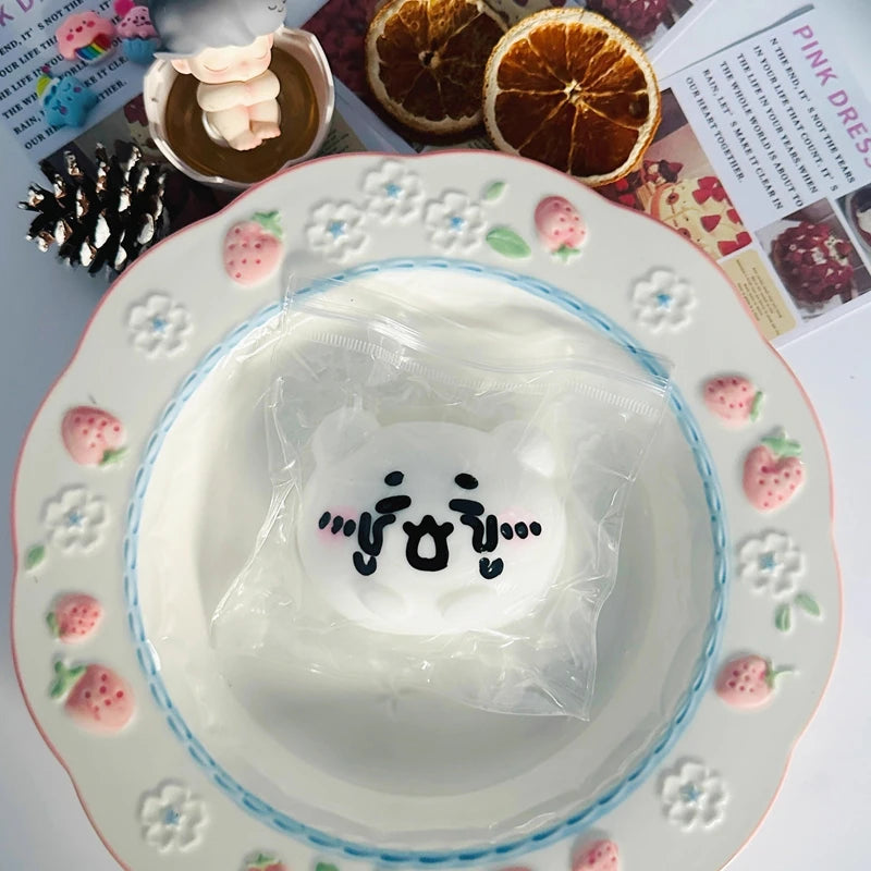 A white squishy toy shaped like a cat face is placed in a decorative plate surrounded by small pink flowers and strawberries.