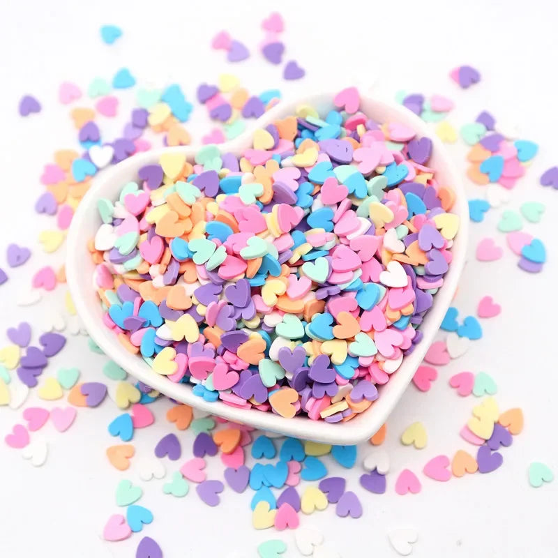 A small white bowl filled with colorful heart-shaped polymer clay sprinkles scattered on a white surface.