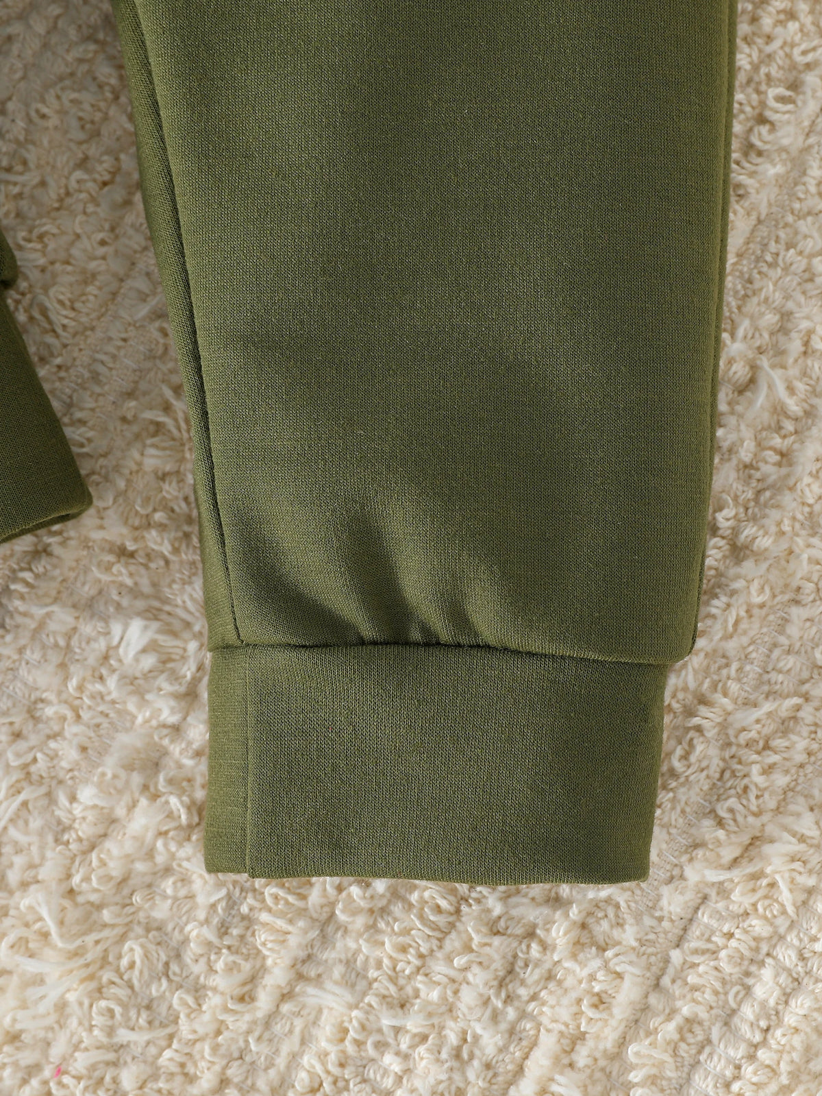 A close-up of a dark olive green boys' sweatshirt sleeve with contrast panels lying on a light beige, fluffy surface.
