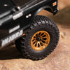 A close-up of a black RC truck wheel with a black beadlock tire and gold accents on a rock surface.