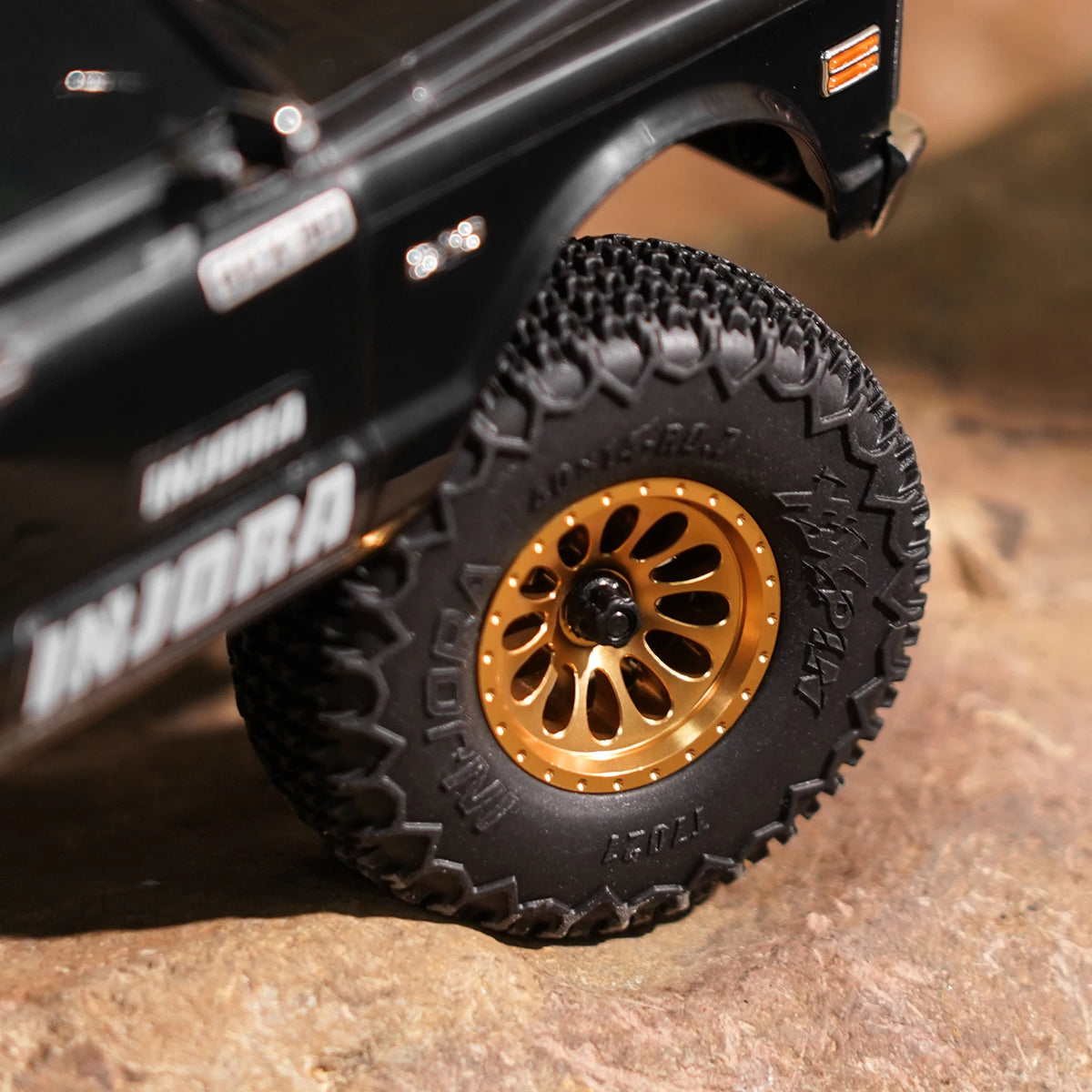 A close-up of a black RC truck wheel with a black beadlock tire and gold accents on a rock surface.