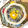 A clear plastic rattle and hand drum set with a yellow tiger design placed on a wooden tray surrounded by small white beads.