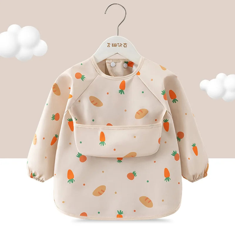 A beige toddler feeding bib with long sleeves and a pocket, featuring scattered illustrations of carrots and other vegetables