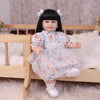 A baby doll with soft cloth body and black hair wearing a floral dress sitting on a wooden bed.